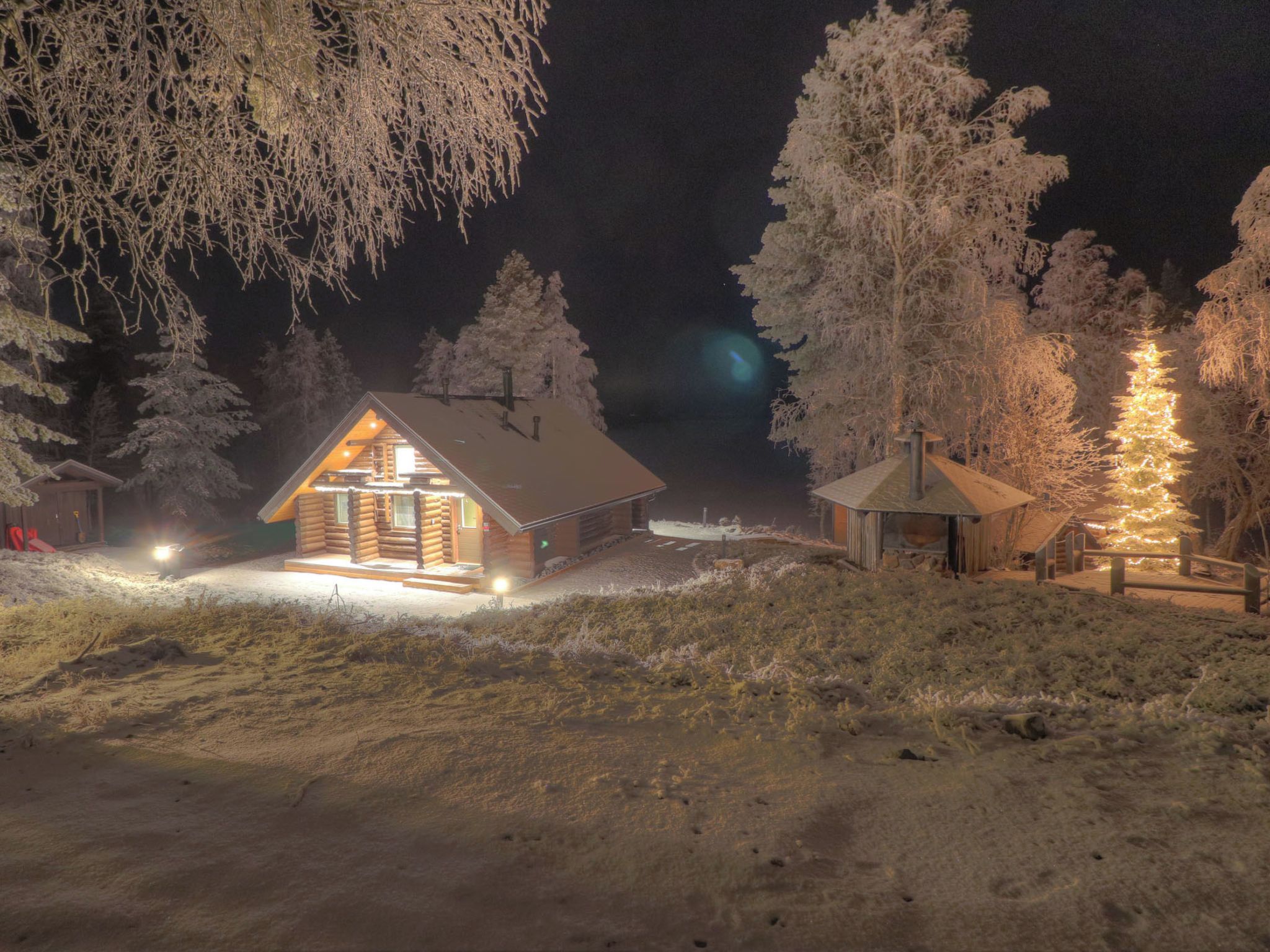 Une cabane en rondins avec un toit en pente, entourée d’arbres couverts de neige et dotée d’une terrasse en bois.