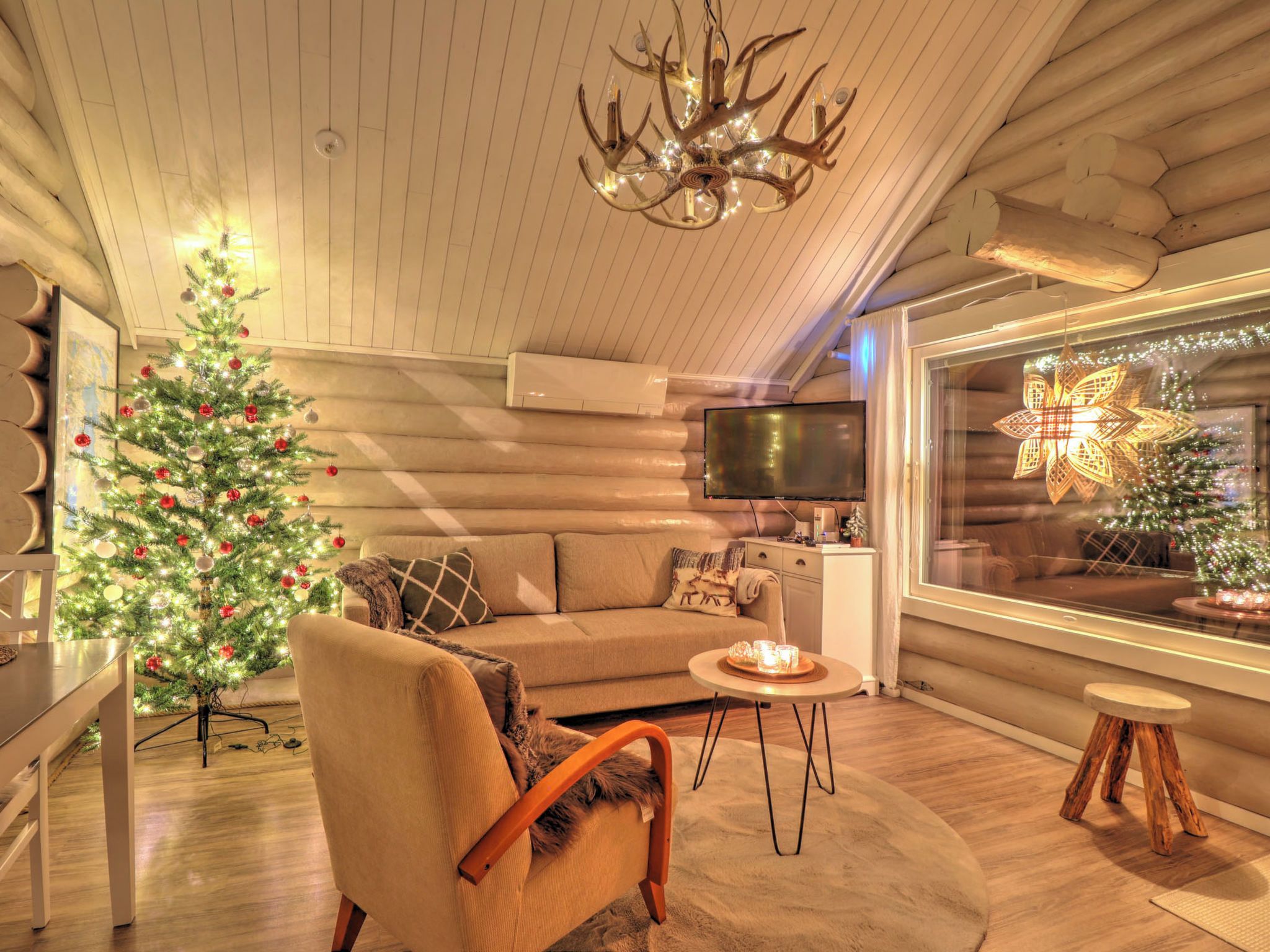 A cozy log cabin interior with a Christmas tree, a chandelier, and a television.