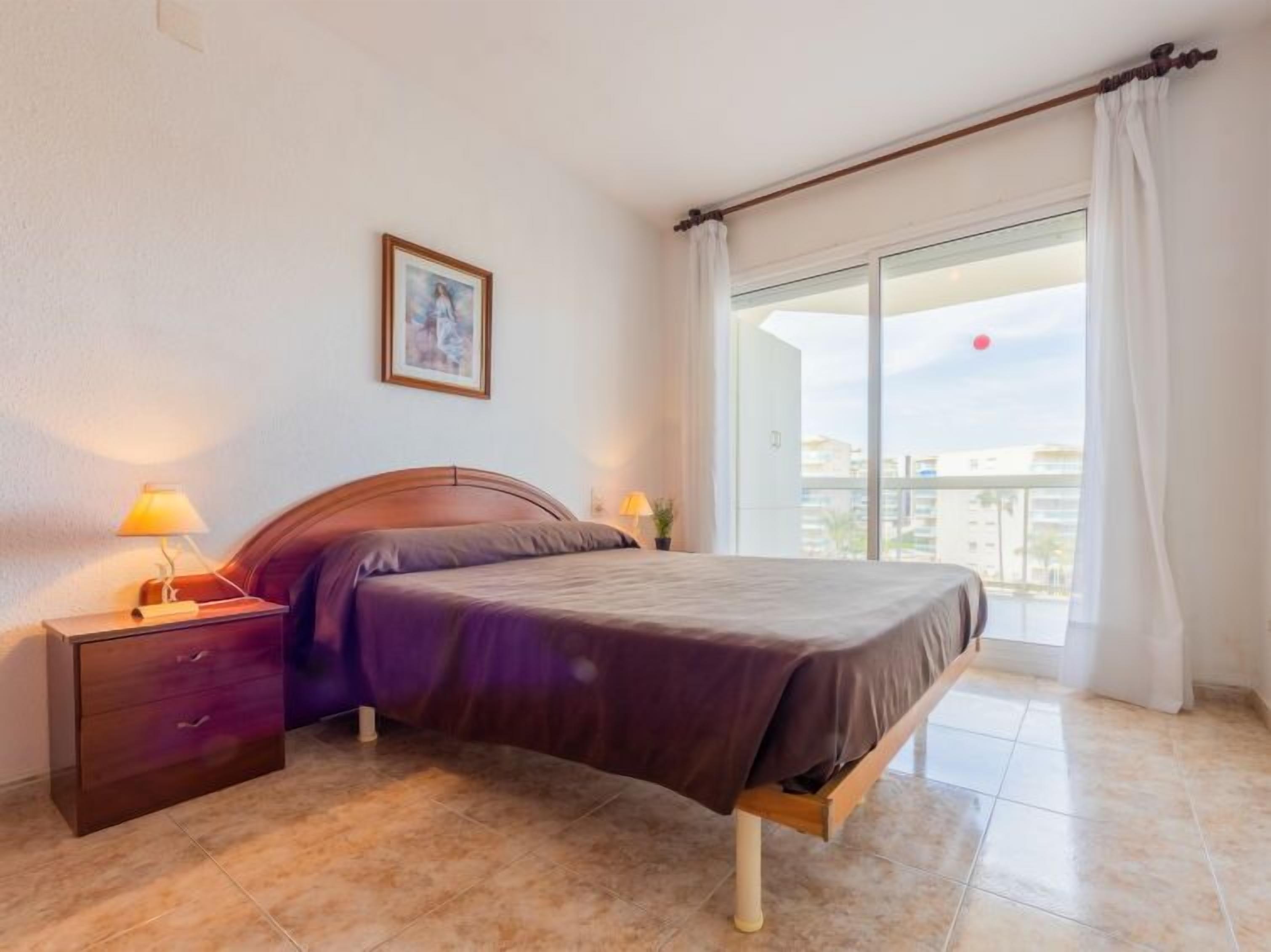 A bedroom with a bed, nightstands, a lamp, a picture on the wall, and a large window with curtains.