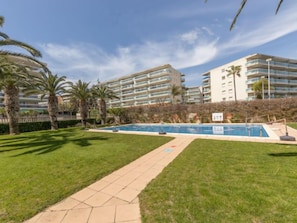 A pool area with a clear sky, palm trees, and a pathway leading to a building with multiple balconies. - Living Park by Interhome (Salou)