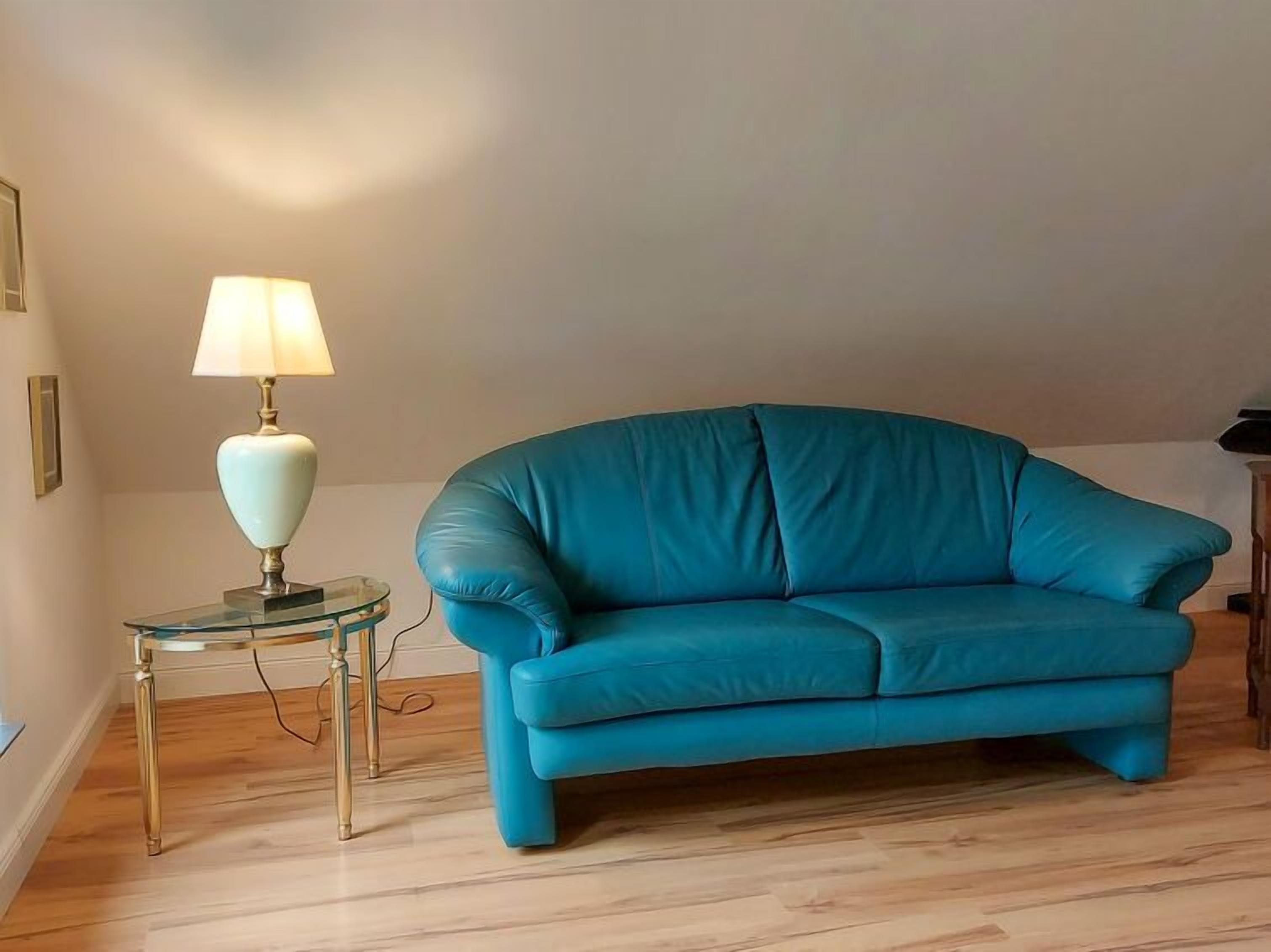 Een smaragdgroene bank in een kamer met houten vloeren en een glazen bijzettafeltje met een lamp.