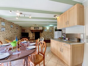 A dining area with a stone wall, wooden chairs, a table set for two, and a kitchen with wooden cabinets and a microwave. - Vacation home Ty Coz by Interhome (Pleurtuit)