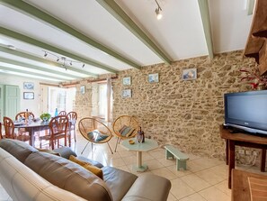 A cozy living room with a stone wall, a sofa, a dining table with chairs, and a television on a wooden stand.