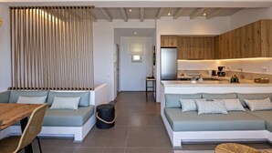 A modern living room with a sofa, a dining table, and a kitchen area with wooden cabinets and a refrigerator. - Villa Erato in Triantaros Tinos (Tinos island)