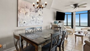A dining area with a large abstract painting, a chandelier, and a view of the ocean through large windows. - Large, oceanfront family condos in quiet, secluded area-balcony, pool (North Myrtle Beach)