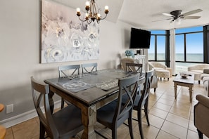 A dining area with a large abstract painting, a chandelier, and a view of the ocean through large windows. - Large, oceanfront family condos in quiet, secluded area-balcony, pool (North Myrtle Beach)