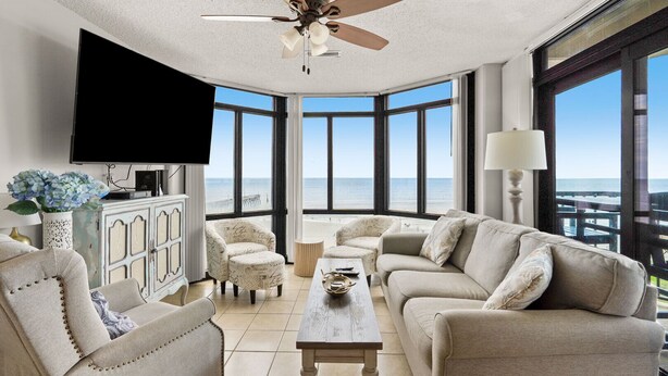 A spacious living room with a large window offering a view of the ocean, a ceiling fan, and a flat-screen TV mounted on the wall. - Large, oceanfront family condos in quiet, secluded area-balcony, pool (North Myrtle Beach)
