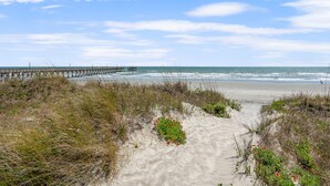 A beach with a wooden pier extending into the ocean, sandy dunes, and sparse vegetation. - Large, oceanfront family condos in quiet, secluded area-balcony, pool (North Myrtle Beach)