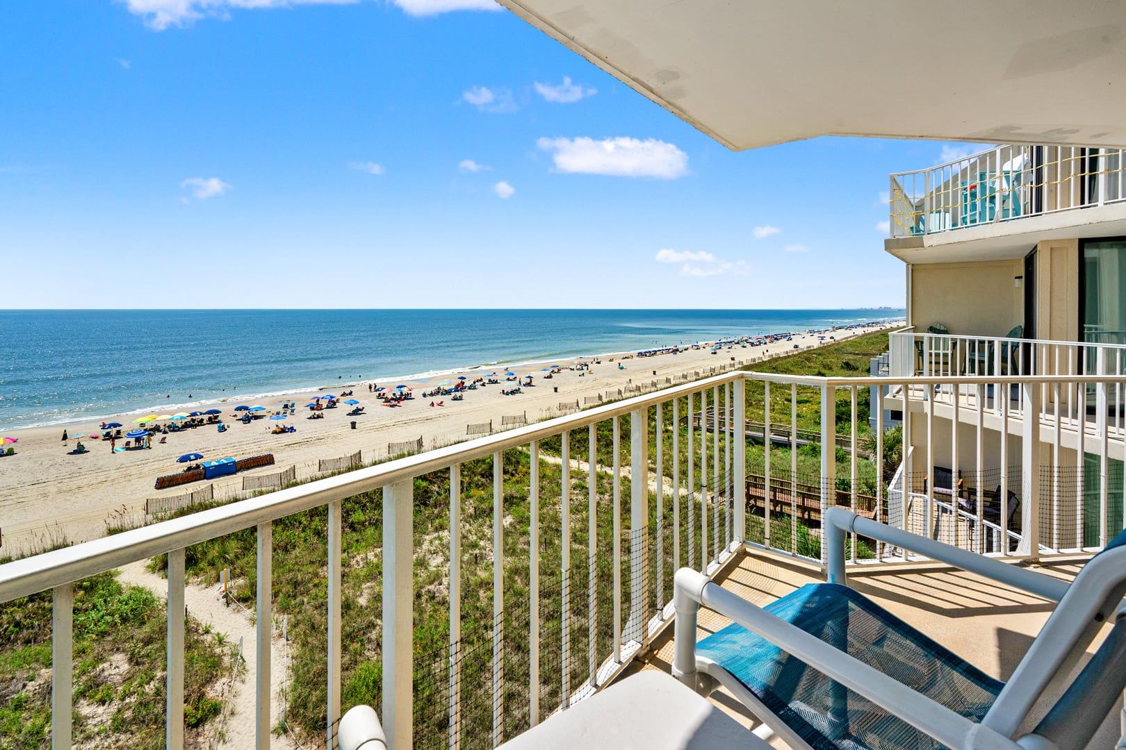 A balcony with a view of a beach, a pool area, and a building with a balcony.