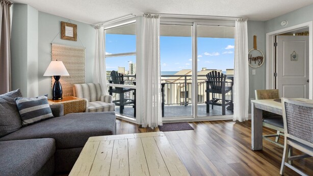 A living room with a sofa, a table, and a lamp. There is a balcony with chairs and a view of the ocean. - Seaside Resort- modern beach destination! (North Myrtle Beach)