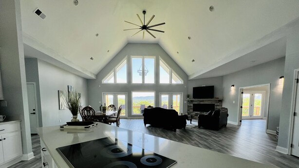 A spacious living area with a large island stove, a dining table with chairs, a sofa, and a fireplace. There are large windows providing natural light and a view of the outdoors. - New! Private 5BR 5,000 SqFt Lake House on 7 Acres (Dandridge)