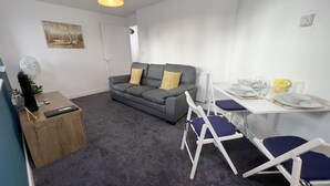 Un espace de vie compact comprenant un canapé gris, deux coussins jaunes, une petite table d’appoint en bois avec un haut-parleur, une table à manger blanche avec des chaises bleues et un radiateur.
