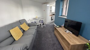 Un salon moderne avec un canapé gris, un meuble TV en bois et un petit coin cuisine à l’arrière-plan.