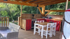 Una terraza exterior con mobiliario de mimbre, una mesa de madera y una zona de cocina con detalles a rayas rojas.