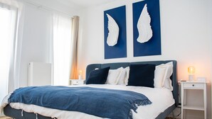 A modern bedroom with a large bed, two wall-mounted artwork pieces, bedside tables with lamps, and a wooden floor.