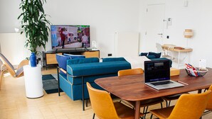 A modern living room with a wooden dining table, yellow chairs, a blue sofa, and a TV displaying a shopping scene.