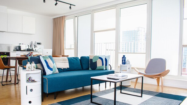 A modern living room with a blue sofa, a coffee table, and a side table. There is a kitchen area with a white countertop and cabinets.