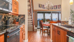 A kitchen with wooden cabinets, a granite countertop, and a view of the ocean through a sliding glass door. - Aloha Condos, Kanaloa at Kona, Condo 2204, Oceanfront, AC (Kailua Kona)