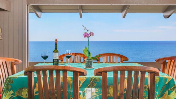 A table set for two with a view of the sea, wooden chairs, and a tablecloth with a tropical leaf pattern. - Aloha Condos, Kanaloa at Kona, Condo 2204, Oceanfront, AC (Kailua Kona)