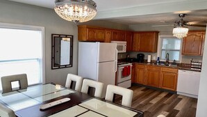 A modern kitchen with wooden cabinets, a refrigerator, microwave, and a dining area with a glass table and chairs. - NEW REMODEL! ARCADE GAMES, PNG PNG, SKEE BALL, ACCESS TO POOL. WE HAVE IT ALL! (Urbandale)