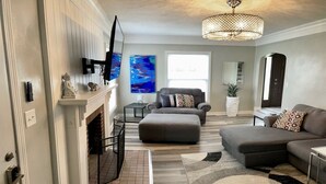 A living room with a fireplace, a large TV, a grey sofa, and a chandelier. - NEW REMODEL! ARCADE GAMES, PNG PNG, SKEE BALL, ACCESS TO POOL. WE HAVE IT ALL! (Urbandale)