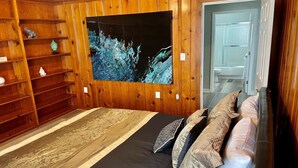 A bedroom with a wooden wall panel, a large abstract painting, a bed with patterned pillows, and a wooden shelf with decorative items. - NEW REMODEL! ARCADE GAMES, PNG PNG, SKEE BALL, ACCESS TO POOL. WE HAVE IT ALL! (Urbandale)