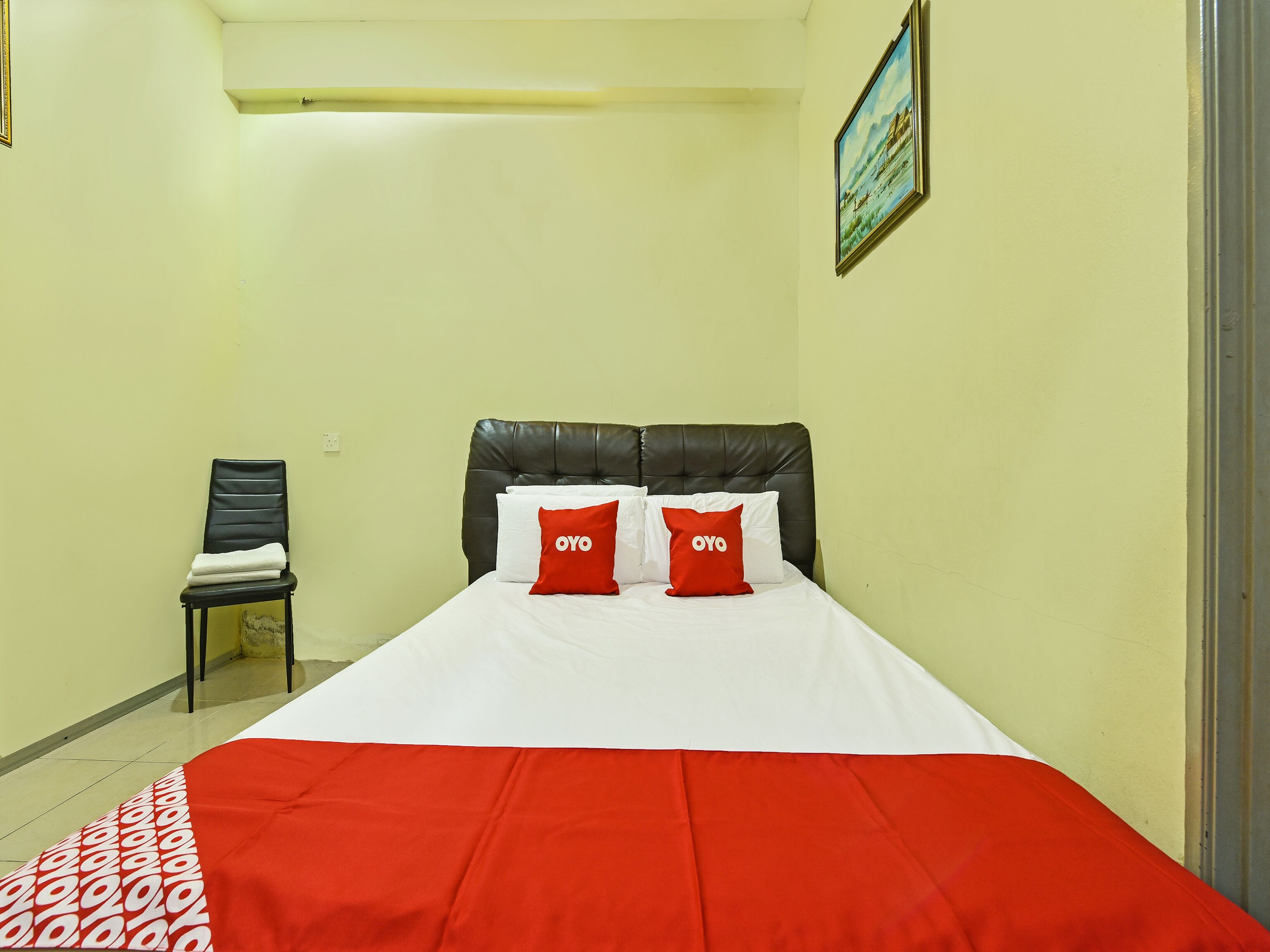 Une chambre d’hôtel avec un lit, deux oreillers rouges portant la marque « OYO », une tête de lit en cuir noir, une couvre-lit blanche et rouge, une chaise noire avec une serviette blanche, et un tableau accroché au mur.