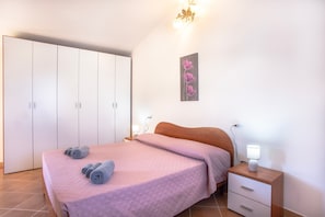 A bedroom with a bed, a nightstand, a wall-mounted lamp, a wardrobe, and a painting. - Villa "Orchidea" with Garden, Terrace, Balcony & Air Conditioning (Masainas)
