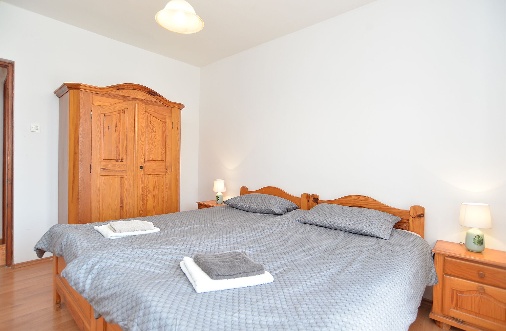 Een slaapkamer met een houten bed, twee kussens, een nachtkastje met een lamp en een houten garderobe.