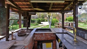 An outdoor patio area with a brick grill, wooden tables, and a view of a garden.