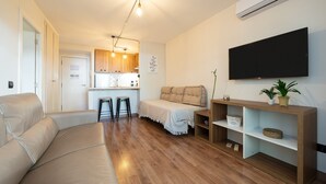 A modern living room with a sofa, a TV, a wooden floor, a kitchen in the background, and a ceiling light. - Apartment 'Apartamentos Salou - 112' with Shared Pool, Wi-Fi and Air Conditioning (Salou)