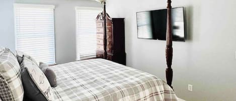 A bedroom with a four-poster bed, a ceiling fan, a flat-screen TV mounted on the wall, and a window with blinds.