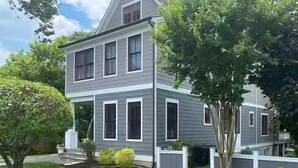 Une maison à deux étages avec un porche à l’avant, un petit jardin et une clôture blanche.
