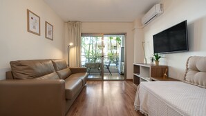 A modern living room with a sofa, a bed, a TV, and a balcony with a view of buildings. - Apartment 'Salou - 111' with Shared Pool, Wi-Fi and Air Conditioning (Salou)
