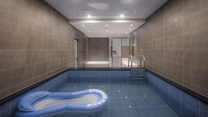 En modern inomhuspool med en blå, uppblåsbar badleksak, en jacuzzi med glasväggar och ett kaklat golv.