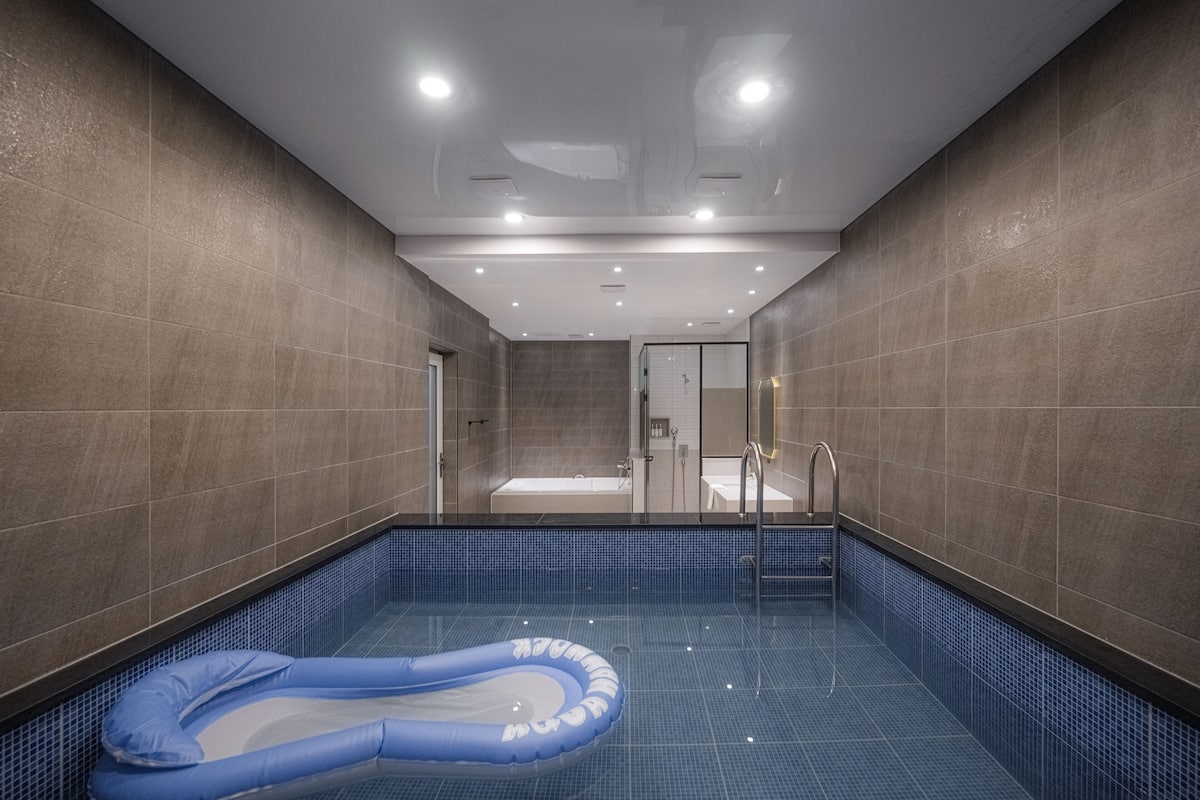 A modern indoor swimming pool with a blue inflatable float, a glass-enclosed jacuzzi, and a tiled floor.
