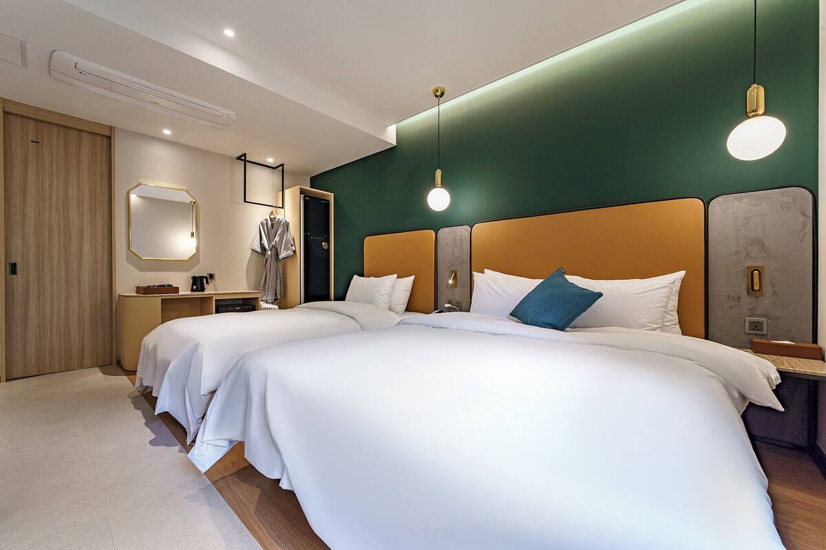 A hotel room with two beds, a wooden wardrobe, a mirror, and a small desk with a lamp.