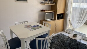 Pieni, hyvin valaistu huone, jossa on valkoinen pöytä, kaksi tuolia, kirjahylly, televisio ja ikkuna, jossa on ohuet verhot.