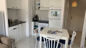Kompakti keittiö, jossa on valkoiset kaapit, sisäänrakennettu pesukone, ruokapöytä tuoleineen ja riippuvalaisin.