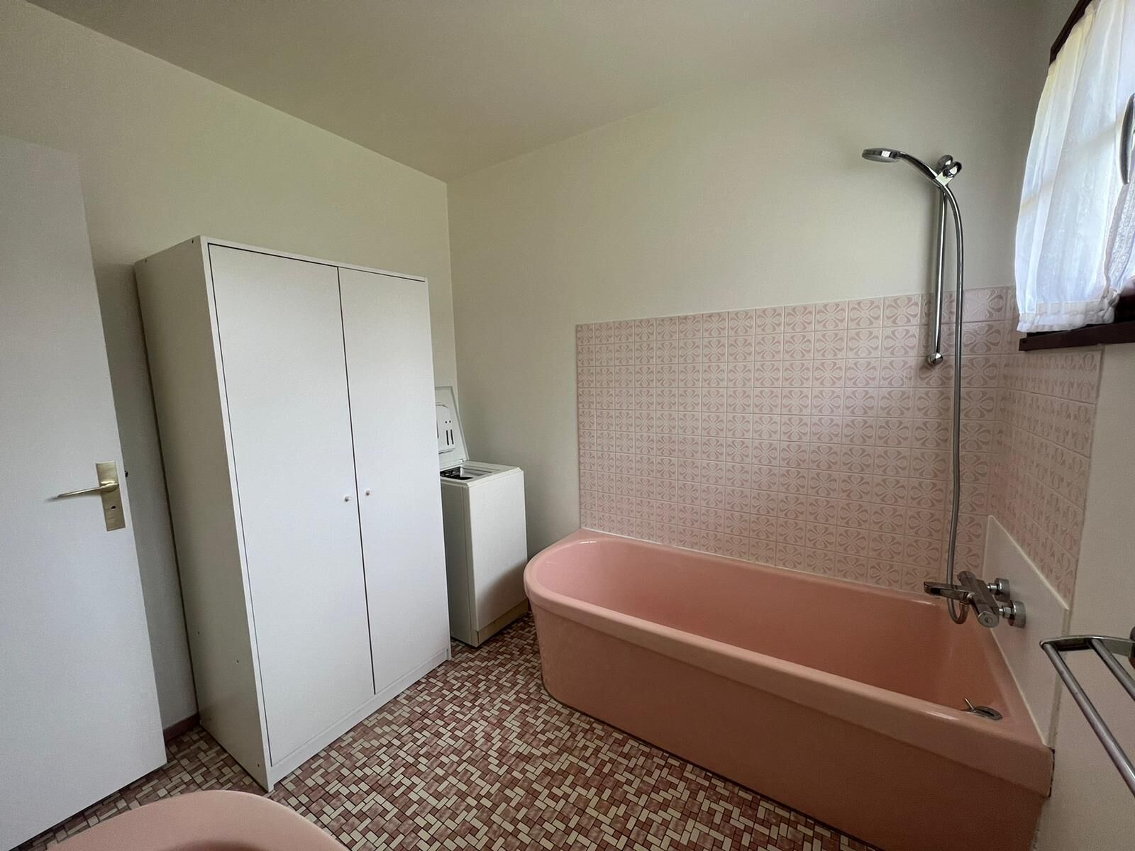 A bathroom with a pink bathtub, a white wardrobe, and patterned floor tiles.