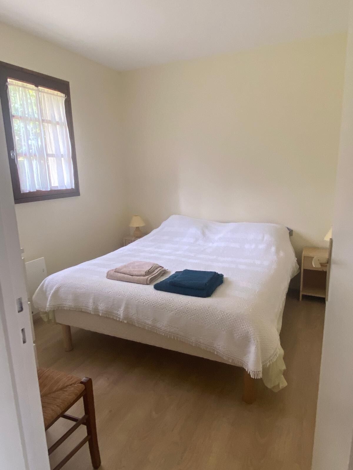 Un dormitorio con cama, una silla, una mesita de noche y una ventana con cortinas.