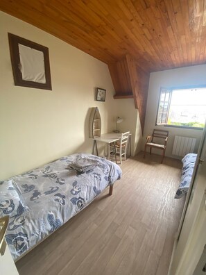 Une petite pièce agréable et chaleureuse, avec un plafond en bois, un lit recouvert d’une courtepointe à motifs, un bureau avec une chaise et une fenêtre donnant sur un paysage verdoyant.