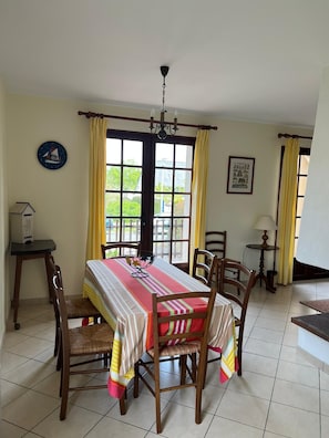 A dining room with a table set for four, surrounded by wooden chairs. There is a window with yellow curtains, a wall clock, and a small table with a lamp.