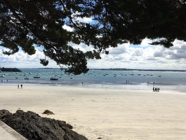 A beach with boats in the water, people walking, and a rocky area in the foreground.