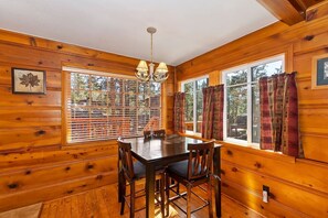 A wooden cabin interior with a dining table and chairs, large windows with curtains, and a chandelier.