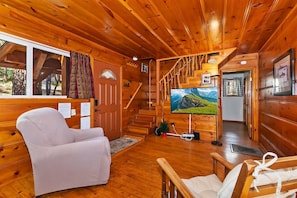 Interior de una cabaña de madera con un sofá, una silla, un televisor y una escalera.