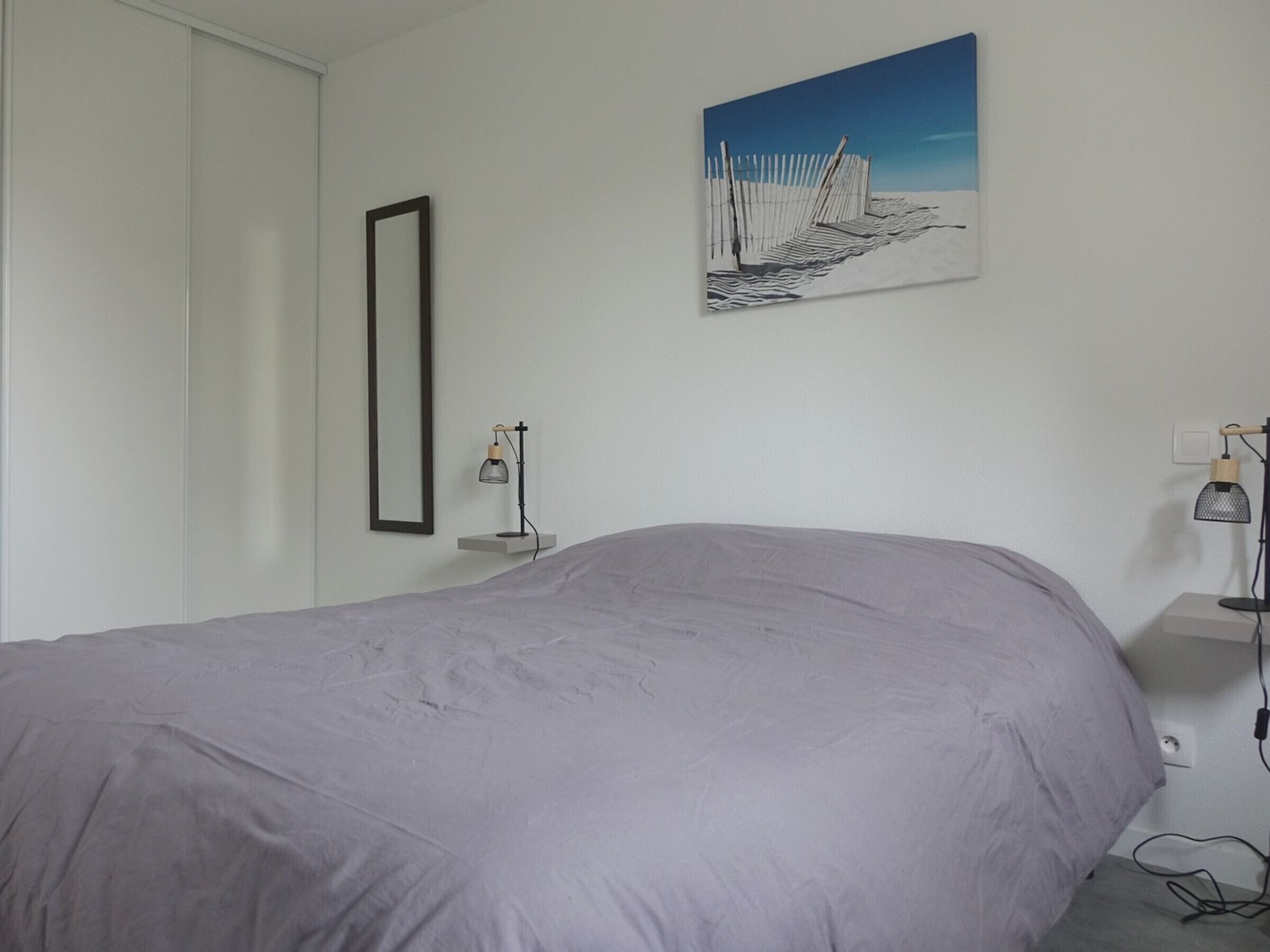 A bedroom with a large bed, a wall-mounted picture, a bedside table with a lamp, and a power outlet on the wall.