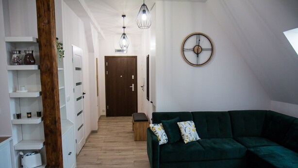 A modern living room with a green sectional sofa, a wooden floor, white built-in shelving, and a wooden door at the end of the hallway. - Apartament Uleczka (Torun)