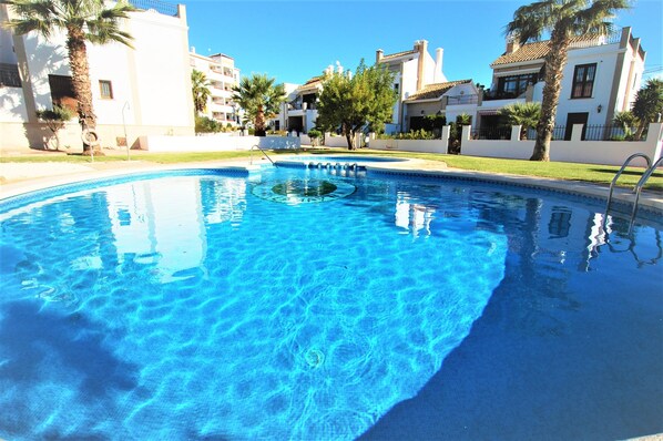 A swimming pool with clear blue water, surrounded by white buildings with balconies and palm trees.