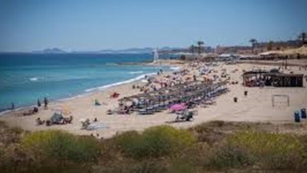 A beach with people and umbrellas, a clear sky, and distant mountains.
